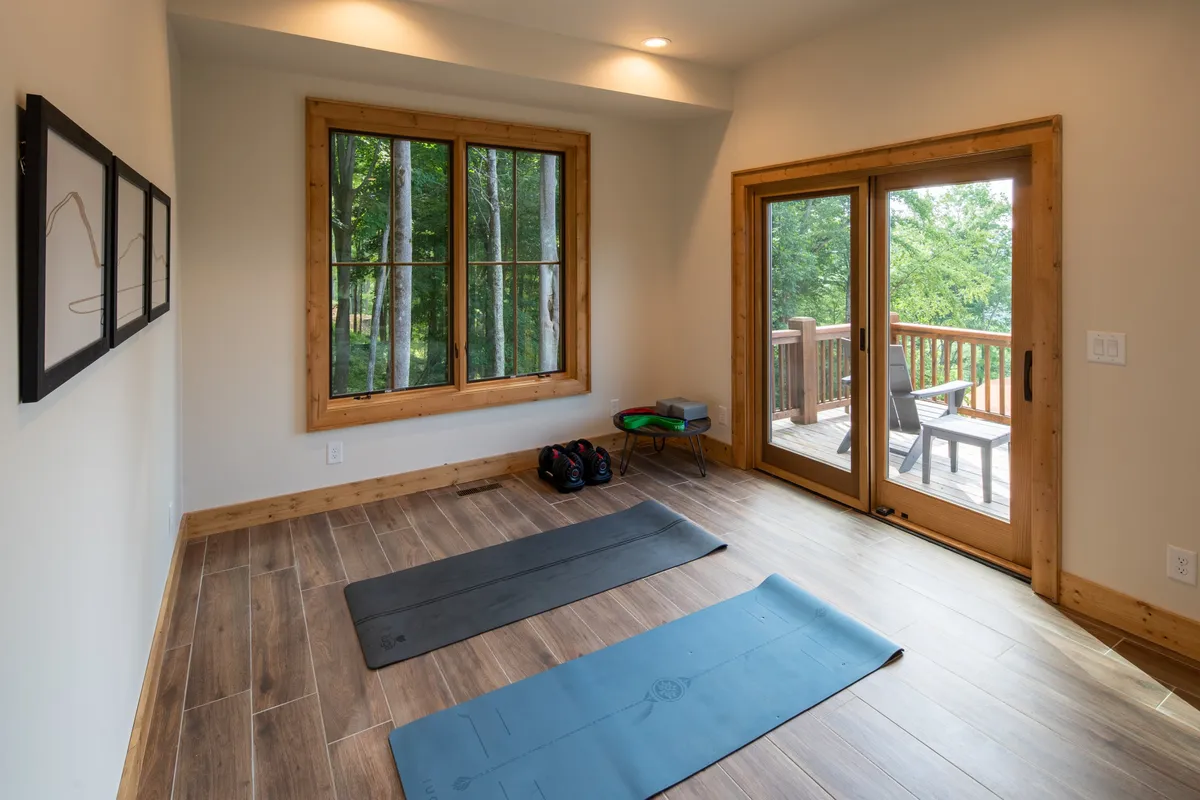 A peaceful fitness and wellness area featuring yoga mats spread across the floor, large windows that flood the room with natural light, and a view of a wooden deck just outside. The tranquil mood sets the scene for a quick workout or a meditative session before heading out for outdoor adventures.