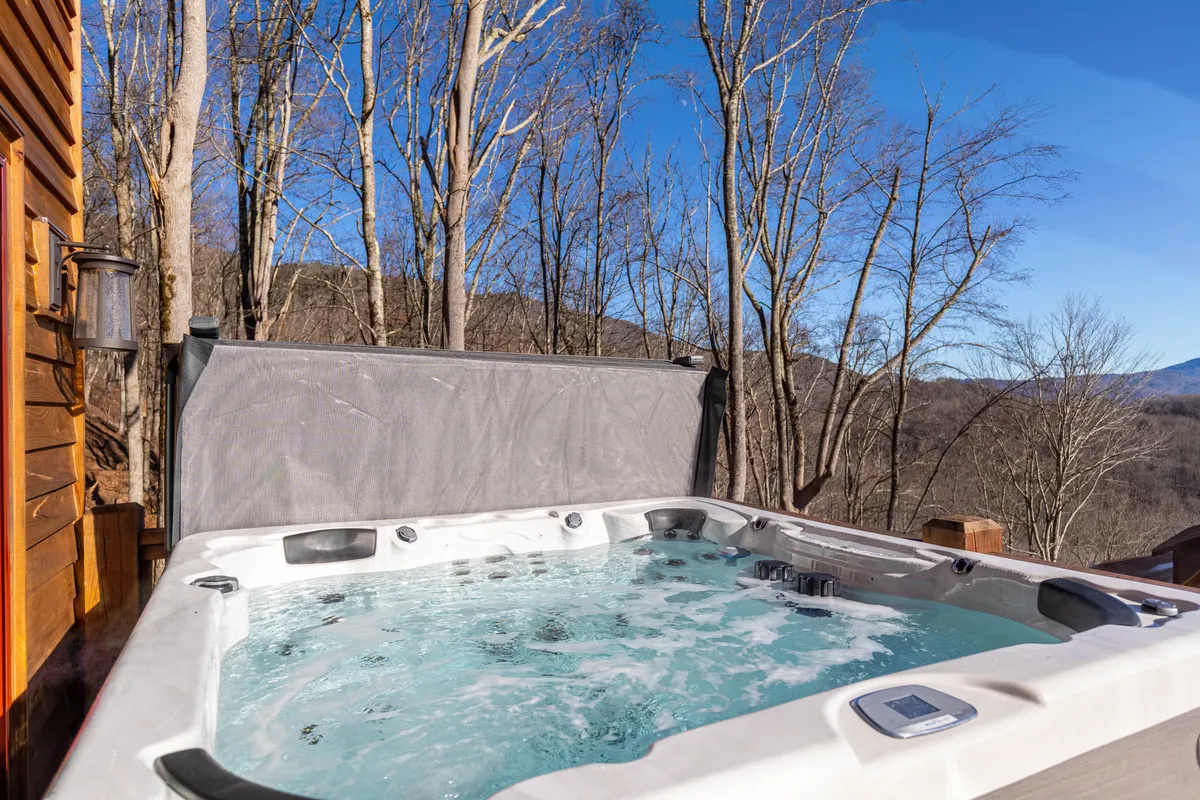 A private hot tub set on a deck, surrounded by lush trees with a sweeping mountain view in the distance. The setting is tranquil, perfect for relaxing outdoors while immersed in nature. The hot tub area is ideal for unwinding with friends after a day of activities or enjoying a celebratory evening under the stars.