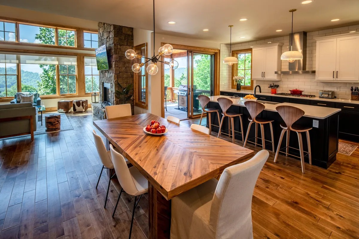 A spacious, sunlit great room featuring a large wall of windows with breathtaking mountain views. The room centers on a striking stacked stone fireplace, with a cozy, inviting seating area perfect for family gatherings or relaxing by the fire. The adjoining kitchen island with bar stools and a rustic wooden dining table are visible, creating a seamless flow between living, dining, and culinary spaces.