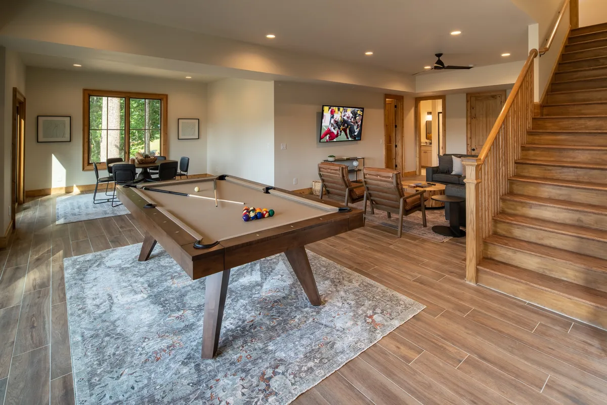 A vibrant interior view of the game room, featuring a pool table as the central focus. Surrounding the table is a cozy seating area, a large television mounted on the wall, and a visible staircase that connects this entertainment space to the rest of the home. The overall atmosphere is casual and inviting, ideal for friendly competition and group gatherings.