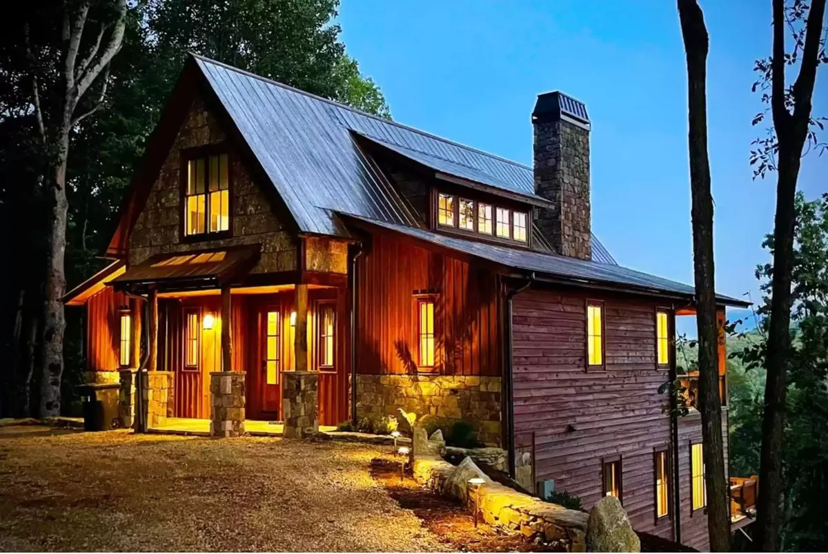 The image features the inviting exterior of a modern mountain home, showcasing a striking stone chimney, a charming wooden facade, and warmly glowing windows nestled among tall trees. The spacious porch offers views of serene nature, perfect for sunset watching, while hints of cozy gathering spaces invite guests to relax and enjoy the tranquil setting.