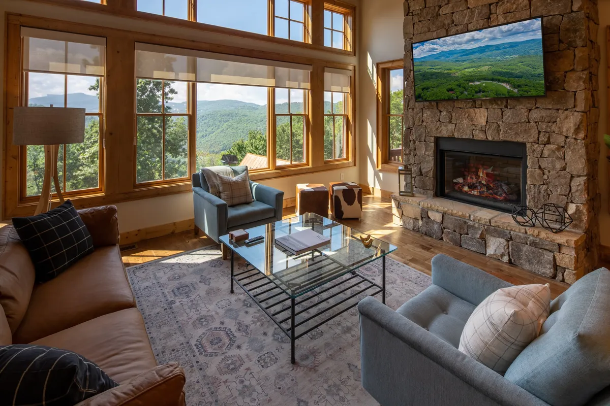 A cozy interior living space featuring large windows that frame expansive mountain views, a stone fireplace, and a comfortable setup for relaxing after a day outside. The scenery through the windows visually connects the interior with the outdoor environment.