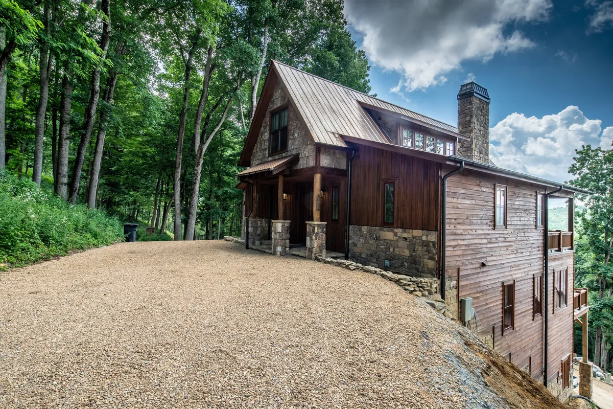 This image showcases the exterior of the property with a wooden and stone facade, a gravel driveway, and surrounding trees that signal instant access to nature. The tranquil scene hints at the adventure possibilities just steps away, with the home nestled seamlessly into the natural landscape.
