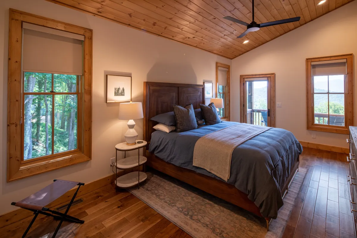 A peaceful bedroom scene with a well-made bed, windows allowing in daylight, a nightstand, and wall art, all arranged to create a calm and inviting environment. The look is simple yet elegant, underscoring comfort and style.