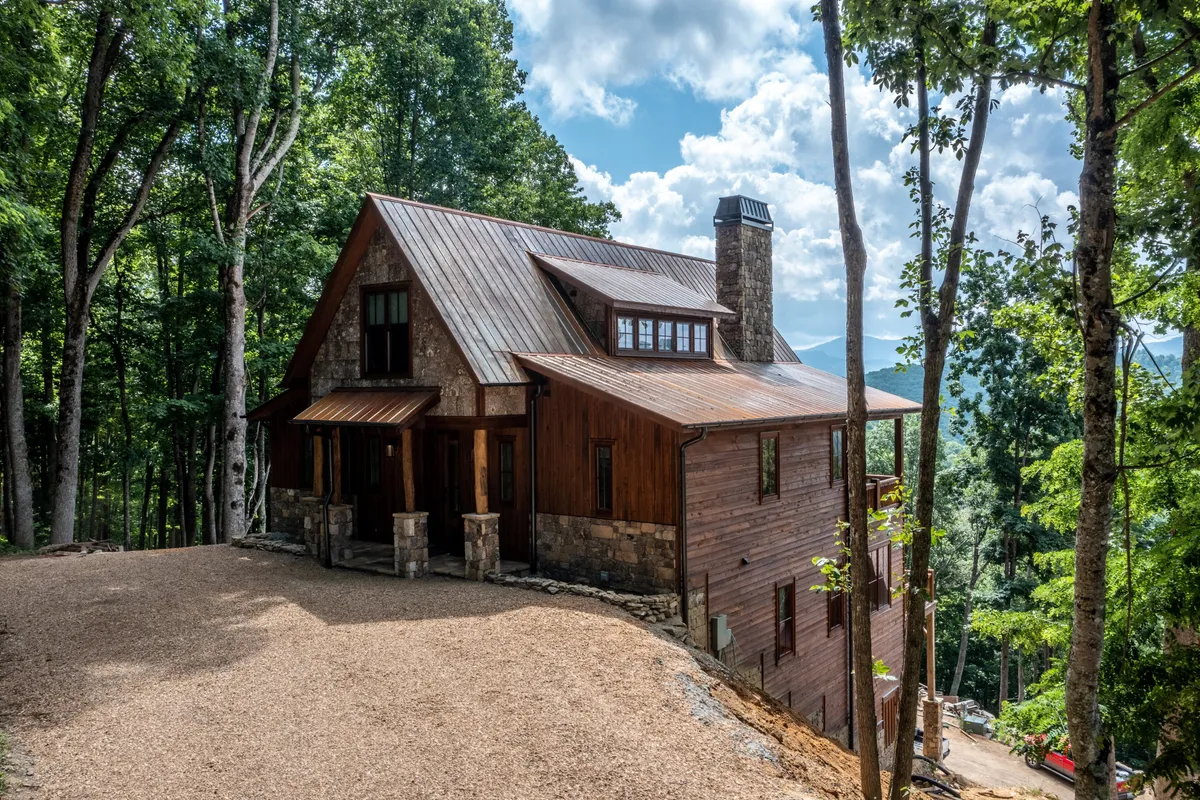 This exterior image highlights the wooden and stone exterior of the home, with a welcoming porch, chimney, and a stunning mountain view in the background. The surrounding trees and natural landscape emphasize the home's connection to its beautiful environment.